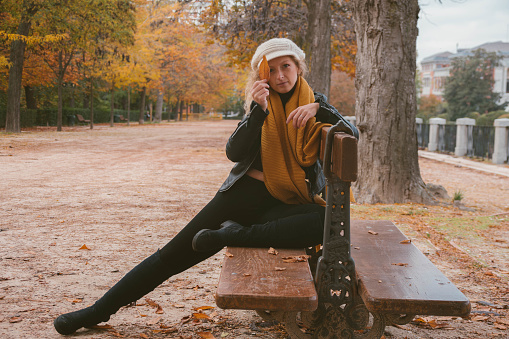 Blonde girl with blue eyes. Dressed in black, with a white hat and an orange scarf. Sitting on a bench with leg stretched. In a park in autumn.