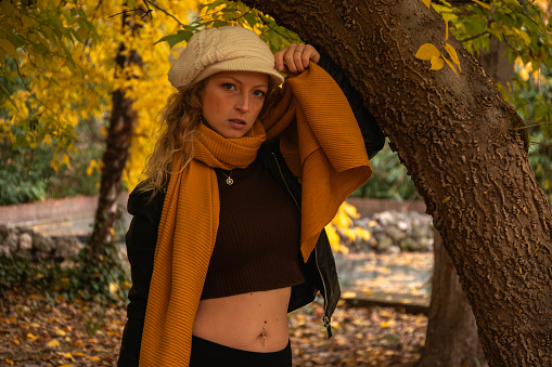 Blonde girl with blue eyes. Dressed in black, with a white hat and orange scarf. Naked gut. In front of a tree. In a park, in autumn. Autumn scene. Half body.