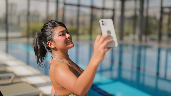 A multiracial female tourist is relaxing and using her mobile smart phone to take photos and videos by the pool during her travel.