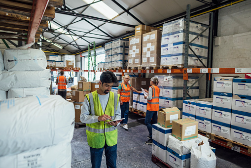 A medium shot of a man and his colleagues in the background wearing casual clothing and reflective vests in an ink factory in Hexham, Northeastern England. He stands and checks the inventory using a digital tablet.