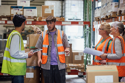 A team of four colleagues wearing casual clothing and reflective vests in an ink factory in Hexham, Northeastern England. They stand and talk with paperwork and digital tablets in hand as they have a meeting.