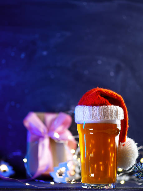 istock A beer glass in a Santa hat on the table with a gift box. dark blue background, copy space 1824638440