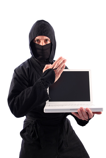 This color photo shows a ninja martial artist dressed in black, guarding a white laptop computer and threatening a knife-hand technique. The laptop computer has a blank screen. The ninja warrior is visible from mid-thigh to the top of his head. The image is isolated on a white background.