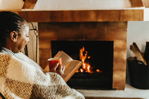 Senior African woman reading a book while warming by cozy fireplace in winter day. Relax and leisure activities concept
