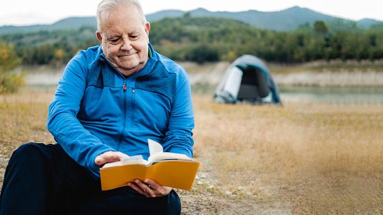 Happy senior man reading a book outdoors during hiking day. Adventure and travel vacations concept