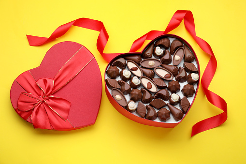 Heart shaped box with delicious chocolate candies and ribbon on yellow background, flat lay
