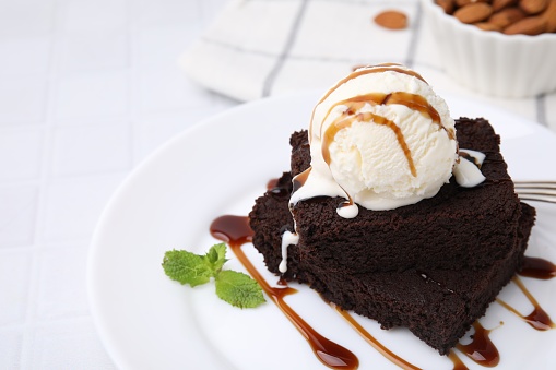 Tasty brownies served with ice cream and caramel sauce on white tiled table, closeup. Space for text