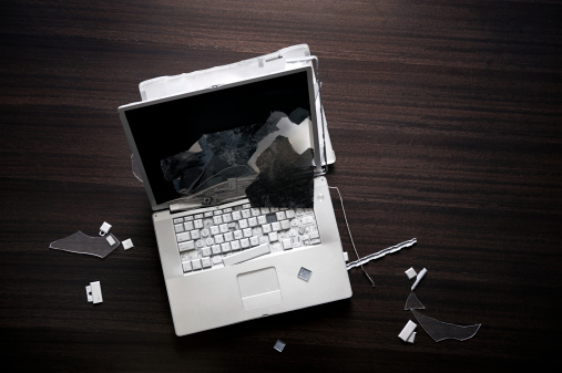 A destroyed laptop on a desk. The screen is smashed and computer keys are torn off, it's sitting on a brown desk. 