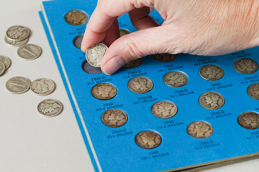 Hand putting an old Mercury Head Dime into a coin collector book.
