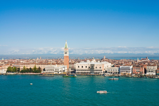 View from the bell tower of San Giorgio Maggiore across the turquoise waters of the Venetian Lagoon towards the landmarks and iconic skyline of Venice, from the mouth of the Grand Canal, past the gondolas and boats, Libreria Sansoviniana and busy Molo waterfront, the distinctive brick tower of the Campanile in St. Mark's Square and along the Riva degli Schiavoni, Doge's Palace and hotels overlooked by the distant snow capped peaks of the Alps on the horizon. ProPhoto RGB profile for maximum color fidelity and gamut.