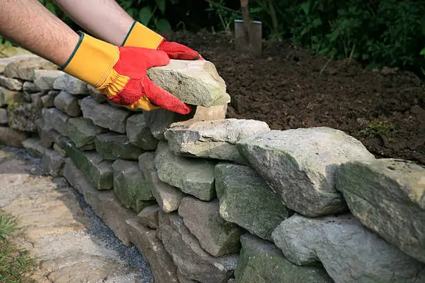 A man with protecting gloves building a drystone wall in the garden.