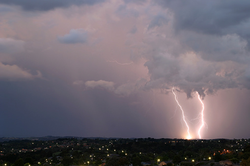 Two cloud to ground lightning bolts strike the ground simultaneously. One of the bolts strikes the power grid, resulting in a bright flash on the ground. In the foreground suburban lights twinkle at night as the storm passes by.