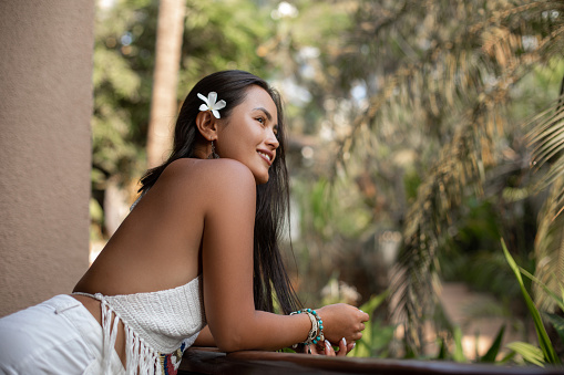 Side view of young attractive Asian woman with dark hair decorated with white orchid leaning on balcony railing, posing.