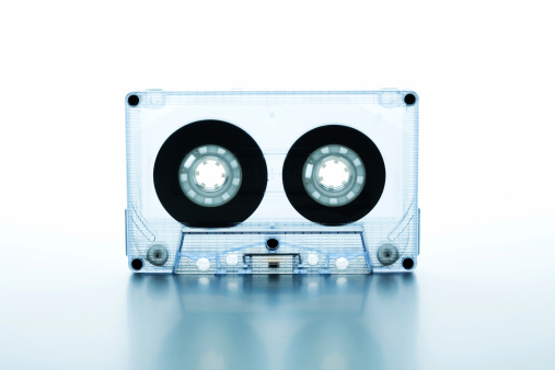 Close up of a clear compact cassette standing on a reflective surface. Studio shot with white background.