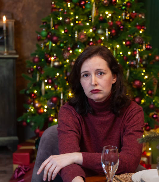 woman sits sadly at table against of decorated Christmas tree stock photo