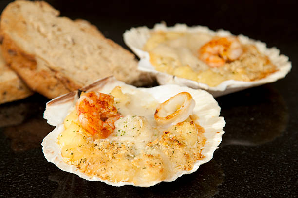 Seafood gratin in scallop shells with scallops, king prawns and squid in a cheese sauce covered with breadcrumbs on a reflective black granite worktop