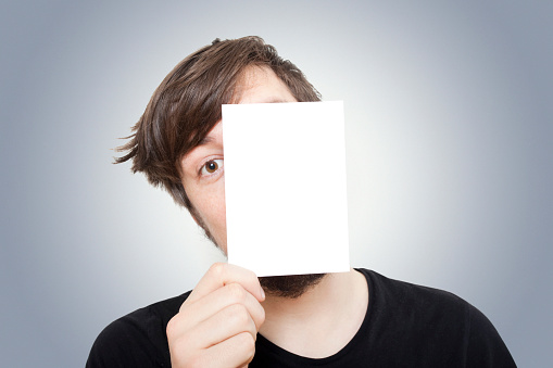 Young man peeping out from behind a paper square which he's holding up in front of his face.Similar images: