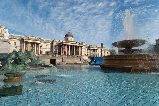 The National Gallery, and fountains, at Trafalgar Square in London. A sunny day, with some light clouds.