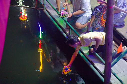 Thai man placing flower bouquet with candles  onto water  at Loy Krathong. Scene is  at canal in Bangkok Ladprao near Wanghin Rd. Night and high angle shot of local Loy Krathong festival. People are  praying  at canal  aopposite  local temple Wat Siri Kamala Vas (Wat Senanikom). There are several people near water