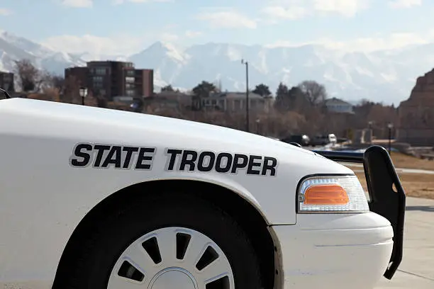 DSLR picture of a white state trooper police car in front of an office building. The sky is blue with few clouds and there is city buildings and mountains in the background.