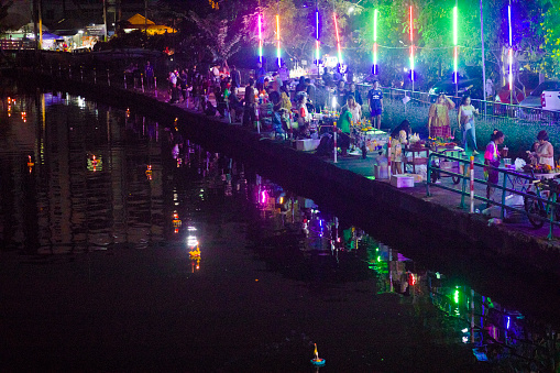 Local Loy Krathong festival at canal in Bangkok Ladprao at night. Neon lights are placed along trees. Lights are multi colored.  Thai people and families are celebrating Loy Krathong at canal in Bangkok Ladprao near Wanghin Rd. People come and go. Some are selling flower bouquets to people.  Scene is opposite to  local temple Wat Siri Kamala Vas (Wat Senanikom). View from footbridge across canal