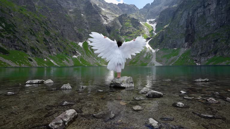 White angel with big wings on a beautiful mountain lake. Young long-haired girl in the image of an angel against a mountain lake and mountains.