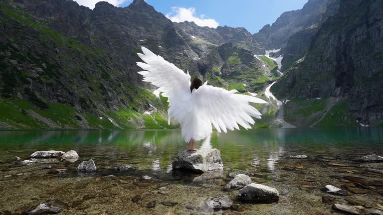 White angel with big wings on a beautiful mountain lake. Young long-haired girl in the image of an angel against a mountain lake and mountains.