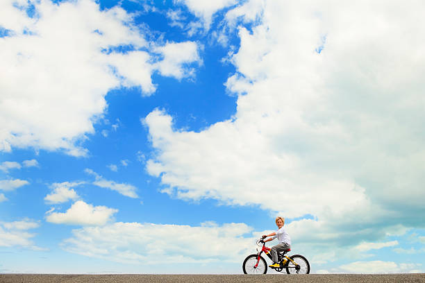 Horizontal image of an 8 year old attractive blond haired boy riding a brightly coloured bicycle confidently along a road on a summer's day. Beautiful blue sky with white clouds behind him. Side view of child but with face turned to camera.