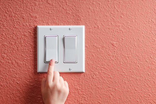 A young finger points upward to click off an unused light.  The light switch is on a sandstone wall with copy space on the right side of the switch.