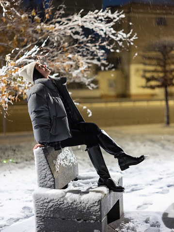 Young woman sitting on bench among snowy winter park at night against background of city lights.