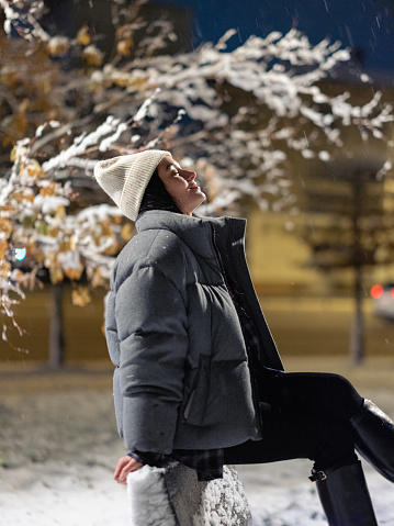 Young woman sitting on bench among snowy winter park at night against background of city lights. Closeup.