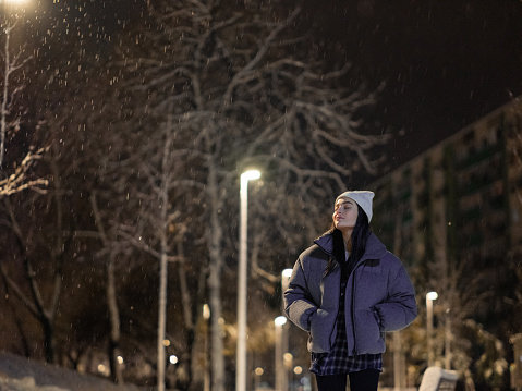 Young woman walking on walkway among snowy winter park at night against background of streetlights.