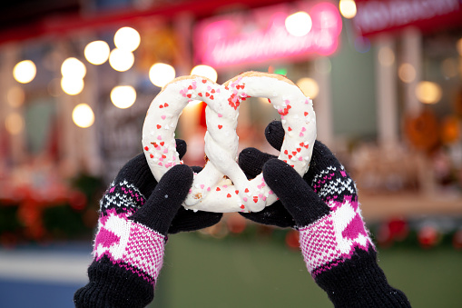 Hands in winter gloves hold a large pretzel in white glaze against the background of colorful street lights