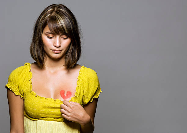 Portrait of young woman with broken heart painted on chest against gray/neutral background with copy space.