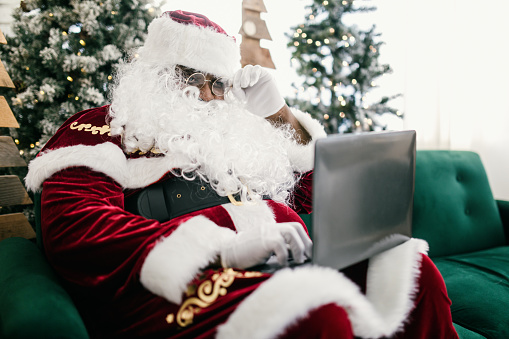 An African American man in Santa costume representing for the Christmas holiday.  He works on a laptop computer, getting last minute Christmas gift requests.  Shot in a festive room with Christmas decorations.
