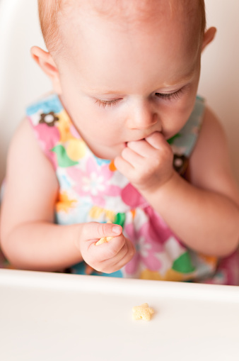 Color photo of a baby girl eating a snack while sitting in a highchair.
