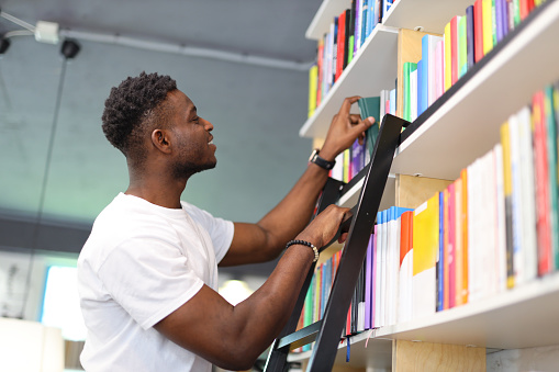 Serious student in a university library, making thoughtful choices from the bookshelf, showcasing intelligence and positivity.