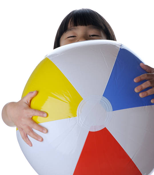 A young girl peeks out from behind a large beach ball she is holding.White background.