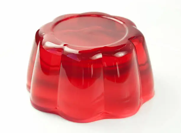 A red piece of gelatin is molded and sitting in the middle of the image over a slightly off-white background.  The light is shining on the mold, casting highlights and shadows.  The mold is shaped like a flower and has smooth sides.