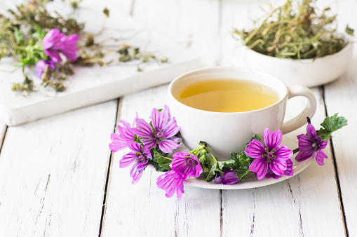 Cup of common mallow tea with fresh blooming malva sylvestris plant on white rustic table, alternative medicine