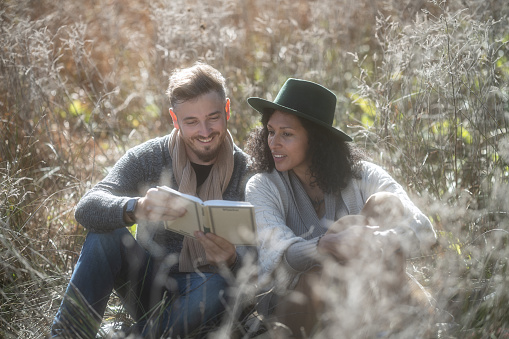 A beautiful Latin American woman and a Caucasian man happy, in love and smiling are sitting on a date in the tall yellow grass in autumn and having fun reading a book. They are leaning on each other. Portrait from the waist up.Sunny weather and morning mists, romance.