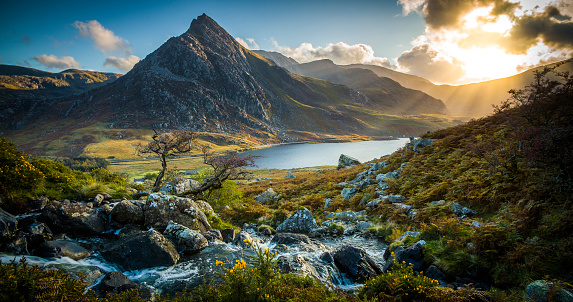 Landscape at Snowdonia National Park in Wales. Mountain stream with fresh water in the foreground. Tryfan mountain and Llyn Ogwen in the background. Warming evening sunlight producing sunbeams in the distance. Northern Wals, UK