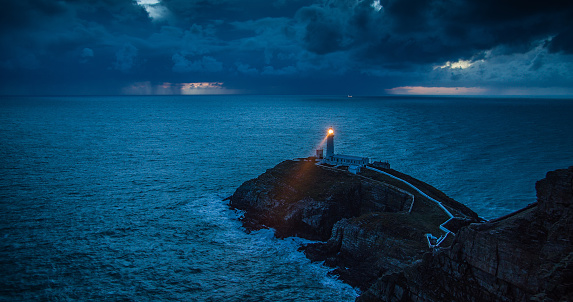 Light beam of the south stack lighthouse, near Holyhead in Wales, moving over the stormy coastline at dusk. Beautiful contrast of the warm light from the lighthouse to the blue-cold surrounding. Wales, UK