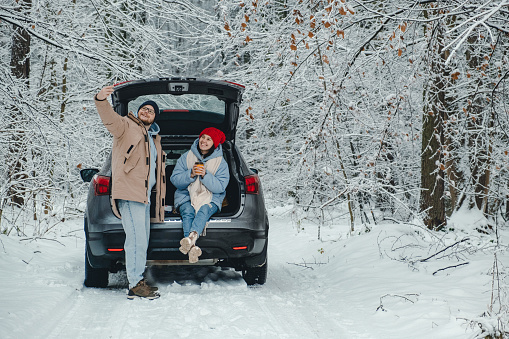 couple takes selfie next to their car in snowy forest when they stop to take a break from the road and eat