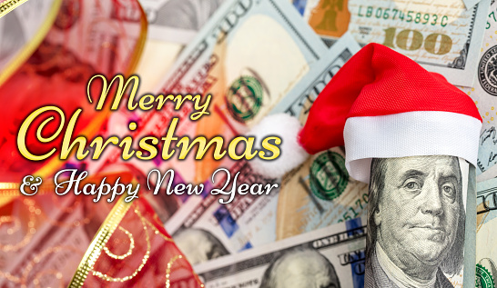 A hundred dollar roll in a Christmas Santa hat as a Christmas present. US 100 dollar bills background. Merry Christmas and Happy New Year lettering.