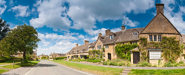 Honey colored limestone homes facing across grassy verges into a quiet road under blue panoramic skies with white fluffy clouds. ProPhoto RGB profile for maximum color fidelity and gamut.