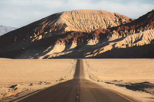 Dramatic golden light on an empty desert road straight ahead through the rugged terrain of the badlands landscape in Death Valley Park National Park, USA.