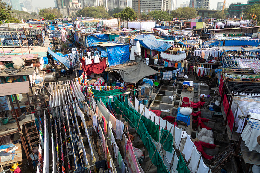 This image captures the bustling activity of the Dhobi Ghat, Mumbai's famous open-air laundromat. Known as the world's largest outdoor laundry, Dhobi Ghat presents a unique aspect of the city's daily life. The photograph showcases rows of concrete wash pens, each fitted with its own flogging stone, where dhobis (washermen and women) vigorously wash a vast array of clothes and linens. The vibrant scene is filled with lines of laundry drying under the sun, creating a colorful mosaic that is both functional and visually striking. The image aims to highlight this traditional laundry method, showcasing the hard work and community spirit of the dhobis, while offering a glimpse into a unique, lesser-seen side of urban life in Mumbai.