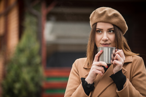 Portrait of a beautiful woman standing next to a wooden log cabin, in nature, drinking hot tea or coffee, enjoying a winter day