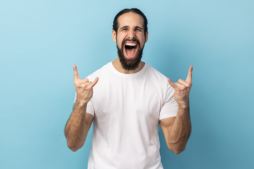 Its rock n roll baby. Portrait of excited positive man with beard wearing white T-shirt looking at camera with rock sing and open mouth. Indoor studio shot isolated on blue background.